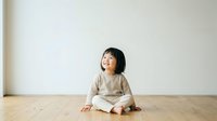 Young Asian toddler in neutral beige clothing sitting on wooden floor against white wall, looking upward with cheerful expression, suitable for parenting and childcare content.