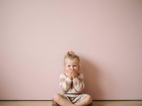 Young blonde toddler girl with fair skin sitting cross-legged against pink wall, covering mouth with both hands in shy or surprised gesture, wearing light sweater. Suitable for parenting, childhood, emotions.
