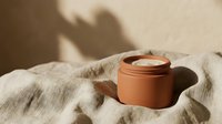 Terracotta clay face mask jar on linen fabric with natural shadow lighting for skincare beauty routine and spa wellness treatment at home.