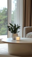 Modern minimalist living room with round coffee table, glowing candle and green plant in white pot near window. Natural daylight creates cozy atmosphere.