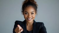 Young African American businesswoman in black blazer pointing forward with confident smile and professional demeanor against gray background. Positive leadership concept.