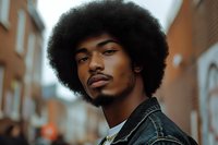 Young African American man with natural afro hairstyle wearing denim jacket poses confidently on urban street, showcasing authentic style and natural beauty in warm evening light.