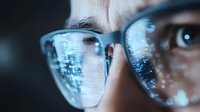 Close up view of eyeglasses reflecting digital data and computer code, showing modern technology interface with glowing blue circuit patterns on dark background.
