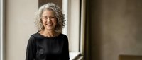 Happy mature Caucasian woman with curly gray hair smiling warmly in black top, standing by window in modern interior for lifestyle and wellness content.