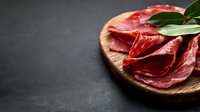 Fresh sliced cured meat on wooden cutting board with bay leaves against dark background, selective focus with copy space for menu design.