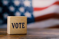 Wooden cube with VOTE text on rustic table against blurred American flag background, representing democracy and electoral process in United States.