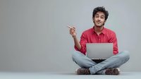 Young man in red shirt sitting cross-legged with laptop, pointing to side with pencil against gray background, suggesting idea or solution.