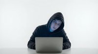 Hooded hacker in dark clothing working on laptop computer for cybersecurity and digital crime concepts in moody lighting against neutral background.