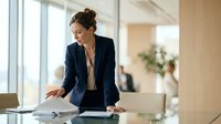 Professional businesswoman in navy suit reviewing documents at modern office desk with natural lighting and contemporary workspace design.