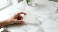 Woman's hand holding blank white business card on marble surface with natural light and green plant, suitable for branding and corporate mockup projects.