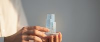 Hands holding transparent glass building blocks creating architectural structure in soft natural light for construction and development concepts.