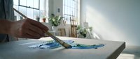 Cropped view of male hand holding paintbrush over watercolor artwork on table in bright studio space with windows and plants.