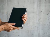 Two hands holding an open black notebook with blank pages against a concrete wall background, suitable for education, business planning, note-taking, and creative projects.