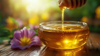 Golden honey drips from wooden dipper into glass bowl surrounded by purple flowers and green foliage, ideal for food, wellness and natural product marketing.