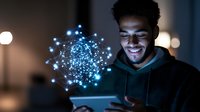 Young African American man in hoodie smiling while using tablet device with glowing blue network hologram visualization floating above screen, dark ambient lighting.