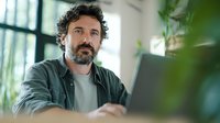 Middle-aged man with curly hair and beard working on laptop in bright home office with plants, representing remote work lifestyle.