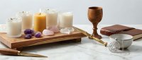 Wooden tray with lit candles, amethyst crystals, wooden goblet, leather journal, and spiritual wellness items on marble surface.