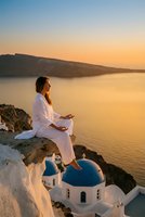 Young woman in white clothing meditating on cliff overlooking sea at golden hour sunset with traditional architecture below for wellness and travel.