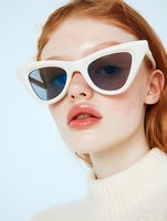 Young redhead woman wearing white cat-eye sunglasses and cream sweater against light background for fashion and eyewear advertising campaigns.