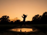 Silhouette of person jumping over water puddle at golden sunset with trees in background, suitable for motivational, lifestyle, and outdoor adventure content.