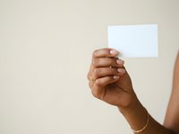 Hand of a person with medium brown skin tone holding a blank white card or paper against a neutral beige background, suitable for business cards, promotional materials, and marketing campaigns.
