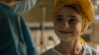 Young Caucasian female patient wearing yellow headscarf smiles optimistically in hospital setting, showing resilience and hope during medical treatment. Warm lighting creates uplifting mood.