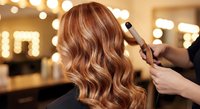Professional hairstylist creating elegant wavy hairstyle with curling iron on woman with auburn hair highlights in modern salon with warm lighting.