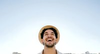 Young man with dark hair and beard, Hispanic ethnicity, wearing tan straw hat and light shirt, smiling outdoors against clear sky for lifestyle and portrait use.