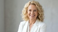 Professional mature blonde businesswoman smiling confidently in white blazer against neutral background for corporate and business presentations.