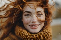 Young redhead woman with freckles smiling genuinely while her curly ginger hair flows in wind, wearing cozy knit turtleneck sweater. Natural autumn portrait outdoors.