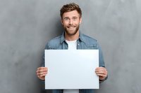 Confident young man with beard holding blank white sign against gray wall background, perfect for adding custom message or promotional content.