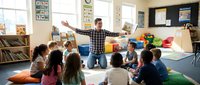 Male teacher in plaid shirt leads interactive lesson with diverse elementary students sitting on colorful floor cushions in bright classroom with educational materials and natural light.