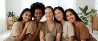 Five diverse women friends embracing and smiling together in cozy earth tone clothing, showing friendship, sisterhood and female bonding in bright indoor setting.