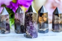 Polished amethyst crystal points with dark inclusions arranged in row on white surface, purple flowers in background create mystical atmosphere.