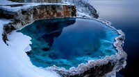 Stunning turquoise hot spring surrounded by snow-covered rocky terrain, creating dramatic contrast between steaming blue water and frozen winter landscape.