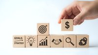 Hand placing dollar sign wooden block completing business strategy concept with goals lightbulb growth handshake search gear icons on white background.