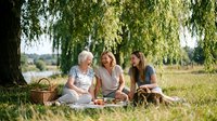 Three generations of women enjoying outdoor picnic under weeping willow tree on sunny day. Grandmother, mother and daughter bonding in natural park setting.