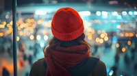Young woman in red beanie and scarf looking at blurred city lights and crowd at night, back view. Atmospheric urban lifestyle scene with bokeh effect and moody colors.