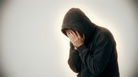 Hooded figure covering face with hands showing despair and emotional distress against neutral background for mental health awareness campaigns.