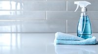Blue spray bottle with cleaning solution and folded microfiber cloth on white surface against tiled wall, home hygiene concept.