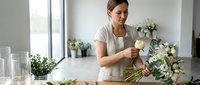 Woman florist arranging white flowers in vase at wooden table in bright studio. Caucasian female professional creating floral arrangement for events, weddings, or retail flower shop business.