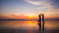 Romantic couple silhouettes holding hands on wet beach sand during golden sunset with colorful sky reflections in water for travel and love concepts.