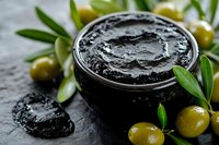 Black olive paste in glass jar surrounded by fresh green olives and olive branches on dark stone background, ready for Mediterranean cuisine.