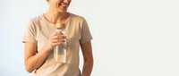 Smiling middle-aged Caucasian woman in beige t-shirt holding glass water bottle on light background, promoting healthy hydration lifestyle.