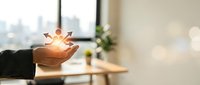 Young professional with dark skin tone holding glowing light bulb with arrows above palm, symbolizing innovation and business growth strategy in modern office workspace.