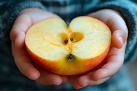 Fresh halved red apple held gently in child's hands against blurred teal background, showing juicy flesh and seeds in natural lighting. Represents healthy eating.