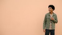 Young man with curly dark hair and medium skin tone wearing olive green shirt over striped undershirt, smiling while gesturing with hand against peachy background, suitable for lifestyle, fashion, and advertising campaigns.