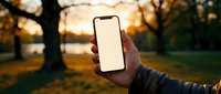Hand holding smartphone with blank white screen in autumn park during golden hour sunset for mobile app mockup and digital technology concepts.