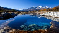 Tranquil mountain lake with crystal clear blue water reflecting snow-capped peaks and autumn foliage in alpine wilderness landscape.