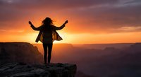 Woman with arms raised celebrating freedom and success on mountain cliff edge during golden sunset with dramatic orange sky and canyon landscape.
